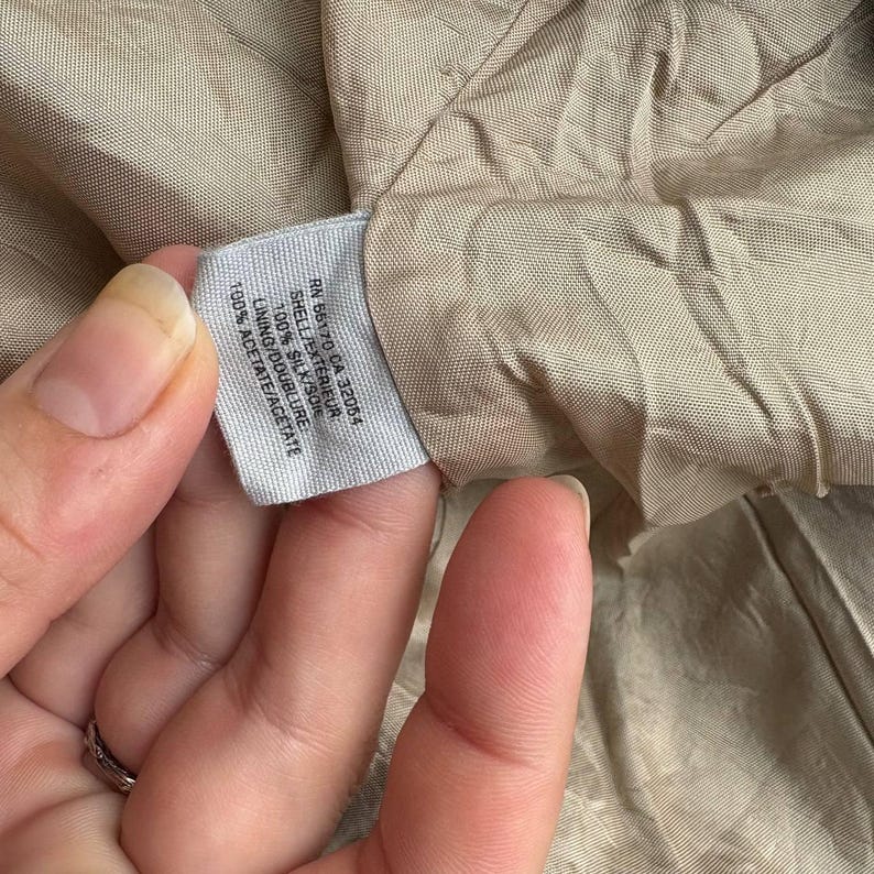May include: Close-up of a tag with text. The tag reads "RN 66170 CA 32054 SHELL/EXTERIEUR 100% SILK/SOIE LINING/DOUBLURE 100% ACETATE/ACETATE". The tag is white with black text. The background is a tan fabric.