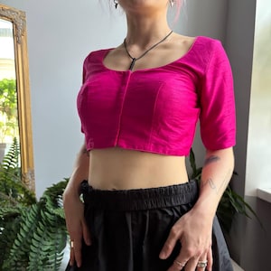 May include: A vibrant fuchsia pink cropped blouse with a scoop neckline and elbow-length sleeves. The garment features a structured bodice with a central vertical seam and a subtle horizontal textured weave throughout the fabric, ending above the waist.
