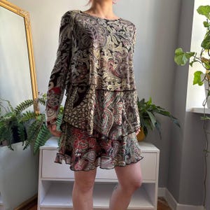 Puede incluir: Un vestido de manga larga con falda de volantes escalonados y cuello redondo. La prenda está decorada con un estampado de cachemira multicolor en tonos negro, crema, rojo y verde, confeccionada con un tejido ligero y semitransparente.