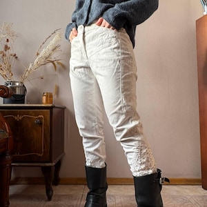 May include: Cream-colored pants with a fitted design and decorative lace detailing at the ankles. The pants have a high waist and a button closure. The model is wearing black boots. The pants are made of a soft, textured fabric.