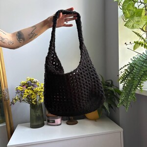 May include: A black crochet shoulder bag featuring an open-knit, textured pattern with a single integrated strap. The bag is constructed from thick yarn, creating a soft, slouchy silhouette with a deep, open main compartment for carrying items.