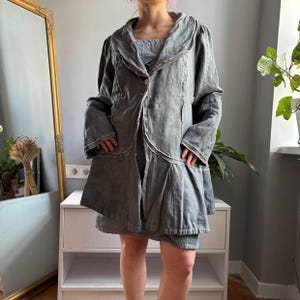 Puede incluir: Una chaqueta de color gris pizarra con mangas largas, cuello ancho, cierre de un solo botón frontal y costuras blancas visibles a lo largo de los bordes. La prenda presenta un dobladillo acampanado y asimétrico con dos bolsillos.