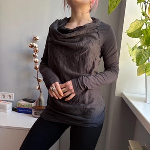 May include: A dark gray long-sleeve shirt with a cowl neck design. The shirt has a textured appearance and is paired with black leggings. The sleeves are long and fitted, and the overall style is casual and comfortable.