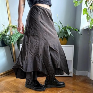 May include: A dark brown midi-length skirt featuring a subtle, tonal swirling circular pattern throughout the fabric. The skirt has a flared, asymmetrical hemline with multiple panels and a smooth, fitted waistband, paired with black leather boots.