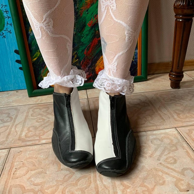 Puede incluir: Botas de tobillo de cuero negro y blanco con una cremallera en la parte delantera. Las botas se llevan con medias de encaje blanco.