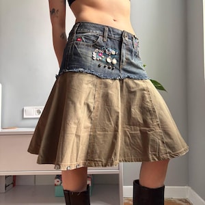 May include: A denim and khaki skirt with decorative embellishments. The skirt features a denim waistband with frayed edges and is adorned with colorful beads and studs. The lower portion is a flared khaki fabric, creating a unique, layered look. The skirt is a mini length.