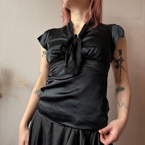 以下が含まれることがあります： 黒いサテンのトップス、首元にリボンディテール。トップスは半袖で、ウエストはギャザー入り。生地はほのかな光沢があり、デザインはクラシックでエレガントです。トップスは黒いスカートと組み合わせています。