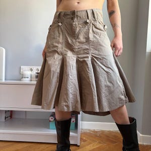 Könnte beinhalten: Ein khakifarbener Rock mit Falten und einem ausgestellten Saum. Der Rock hat einen Knopfverschluss und dekorative Details in der Nähe der Taille. Der Rock wird mit schwarzen Stiefeln kombiniert.