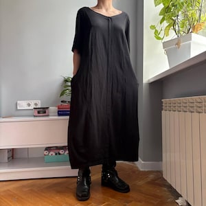 May include: A black, floor-length dress with short sleeves and a V-neck. The dress features pockets and is worn with black boots. The design is simple and minimalist, suitable for casual wear. The dress is approximately 150 cm in length.