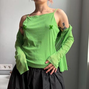 May include: A bright lime green camisole top with thin spaghetti straps, paired with a matching lime green long-sleeved sheer mesh shrug. The camisole features two small green cross-shaped graphics on the chest area, worn over a black skirt.