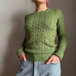 Könnte beinhalten: Ein grüner Zopfstrickpullover mit langen Ärmeln. Der Pullover hat einen Rundhalsausschnitt und gerippte Säume und Bündchen. Die Person trägt eine blaue Jeans. Der Pullover hat eine einfarbige, hellgrüne Farbe.