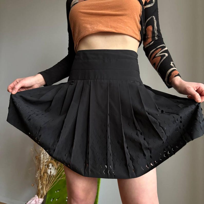 K&ouml;nnte beinhalten: Schwarzer Faltenrock mit hoher Taille und ausgestellter Silhouette. Der Rock hat ein strukturiertes Muster in der N&auml;he des Saums und wird offen gehalten, um seine Form zu zeigen. Der Rock ist aus leichtem Stoff gefertigt.