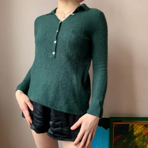 Puede incluir: Un suéter de manga larga verde oscuro con cuello abotonado y un bolsillo en el lado izquierdo. El suéter está hecho de una tela suave y de punto.