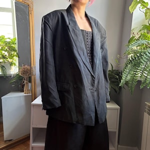 Puede incluir: Un blazer cruzado de color gris carbón con solapa de muesca, mangas largas y dos bolsillos delanteros con solapa. La prenda presenta hombros estructurados y cierre de botones, y se lleva sobre un top oscuro estilo corsé con corchetes.