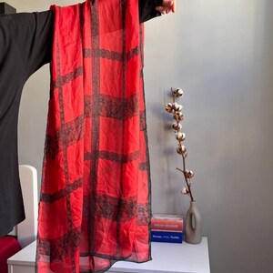 Puede incluir: Bufanda roja transparente con un patr&oacute;n de rejilla de encaje negro. La bufanda se sostiene, mostrando su dise&ntilde;o rectangular y su delicado tejido. Los detalles de encaje a&ntilde;aden un toque de elegancia al accesorio.