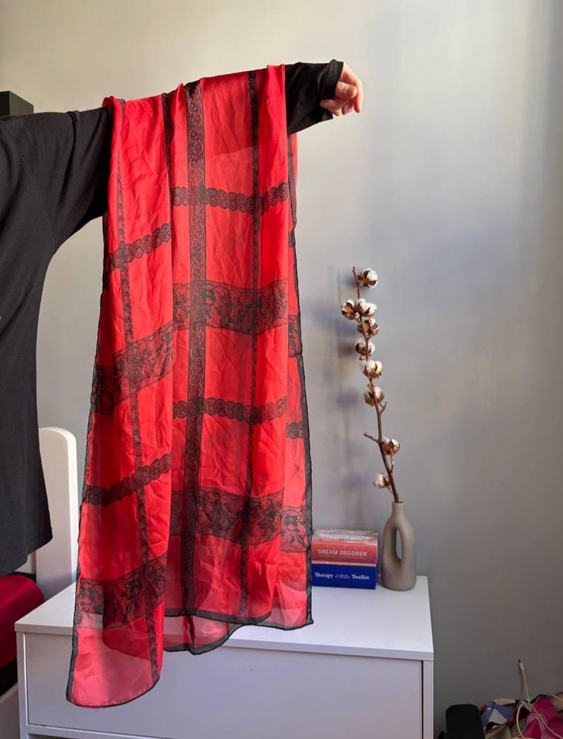 Puede incluir: Bufanda roja transparente con un patr&oacute;n de rejilla de encaje negro. La bufanda se sostiene, mostrando su dise&ntilde;o rectangular y su delicado tejido. Los detalles de encaje a&ntilde;aden un toque de elegancia al accesorio.