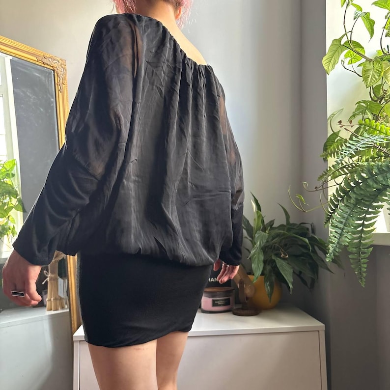 May include: A black long-sleeved top with a loose, sheer, draped bodice and a fitted, opaque lower section. The garment features a wide, rounded neckline and long sleeves, creating a layered appearance with a blouson silhouette at the waist.