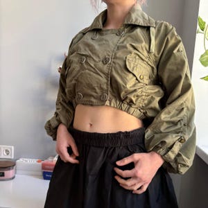 May include: Olive green cropped jacket with double-breasted button closure and two front pockets. The jacket has long sleeves with gathered cuffs. The collar is a classic style. The jacket is made of a lightweight material.