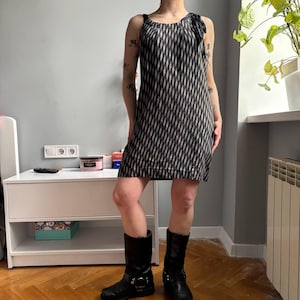May include: A sleeveless black and white patterned dress with a bow detail on the shoulder. The dress is paired with black boots. The dress has a geometric design and is knee-length.