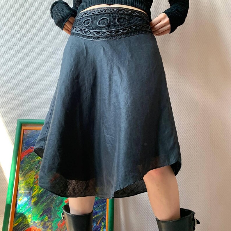 K&ouml;nnte beinhalten: Schwarzer Rock mit dekorativem, besticktem Bund. Der Rock hat eine ausgestellte Silhouette und einen leicht asymmetrischen Saum. Die Person tr&auml;gt schwarze Stiefel. Der Rock ist ca. 60 cm lang.