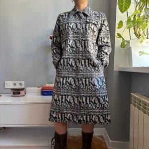 Puede incluir: Vestido negro y blanco con cuello, mangas largas y falda. El vestido está estampado con la palabra "KOOK" repetida en una fuente en negrita. El vestido tiene bolsillos y se usa con botas marrones.