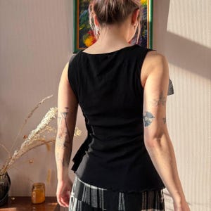 May include: Black sleeveless top with a ruffled side detail. The top has a scoop neckline and a slightly ruffled hem. The fabric appears to be a soft, lightweight material. The person is wearing a black and white striped skirt.