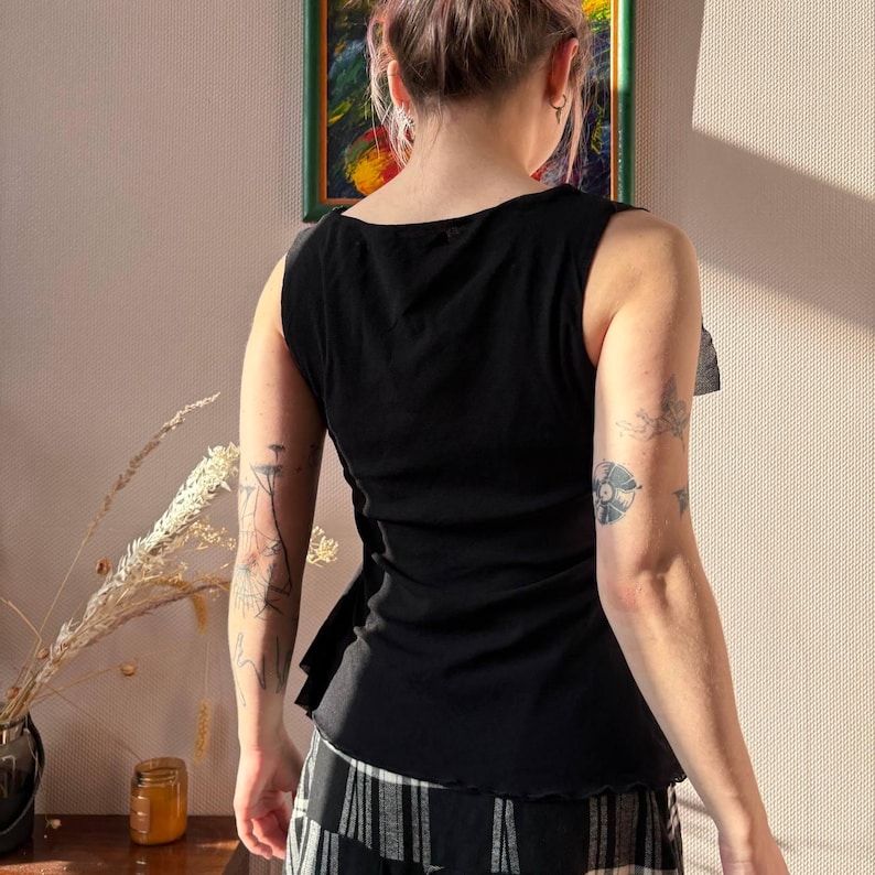 May include: Black sleeveless top with a ruffled side detail. The top has a scoop neckline and a slightly ruffled hem. The fabric appears to be a soft, lightweight material. The person is wearing a black and white striped skirt.