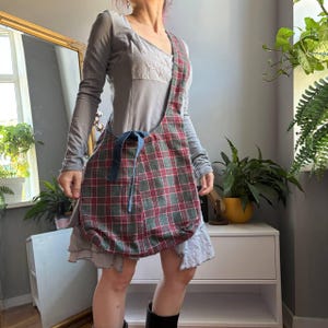 May include: A slouchy hobo-style shoulder bag featuring a red and green plaid pattern. The bag includes a wide integrated shoulder strap and a blue denim fabric tie closure detail on the front, creating a soft, unstructured silhouette.