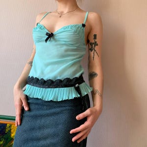 Puede incluir: Camisola azul claro con tirantes finos y lazos negros. La parte superior presenta un diseño plisado con ribete de encaje negro y una banda negra en la cintura. Se combina con una falda vaquera oscura.
