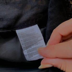 K&ouml;nnte beinhalten: Nahaufnahme eines schwarzen Stoffes mit einem wei&szlig;en Etikett. Das Etikett enth&auml;lt Text in mehreren Sprachen, darunter Deutsch, und Details zur Stoffzusammensetzung und Pflegehinweise. Der Stoff scheint Teil eines Kleidungsst&uuml;cks zu sein.