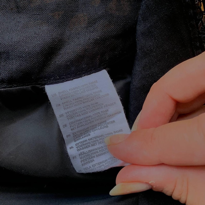 K&ouml;nnte beinhalten: Nahaufnahme eines schwarzen Stoffes mit einem wei&szlig;en Etikett. Das Etikett enth&auml;lt Text in mehreren Sprachen, darunter Deutsch, und Details zur Stoffzusammensetzung und Pflegehinweise. Der Stoff scheint Teil eines Kleidungsst&uuml;cks zu sein.