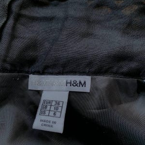 K&ouml;nnte beinhalten: Nahaufnahme eines schwarzen Kleidungsst&uuml;cks mit Etikett. Das Etikett tr&auml;gt den Markennamen "H&M" und Gr&ouml;&szlig;enumrechnungen: EUR 36, GB 10 und US 6. Das Etikett weist auch darauf hin, dass der Artikel in China hergestellt wurde.