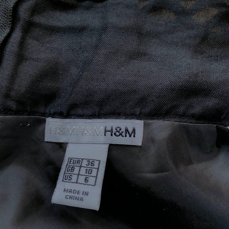 K&ouml;nnte beinhalten: Nahaufnahme eines schwarzen Kleidungsst&uuml;cks mit Etikett. Das Etikett tr&auml;gt den Markennamen "H&M" und Gr&ouml;&szlig;enumrechnungen: EUR 36, GB 10 und US 6. Das Etikett weist auch darauf hin, dass der Artikel in China hergestellt wurde.