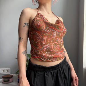 Puede incluir: Top halter con estampado de cachemira en tonos rojos, marrones y blancos. El top tiene un cuello drapeado y está hecho de una tela ligera y transparente. El top se combina con una parte inferior negra.