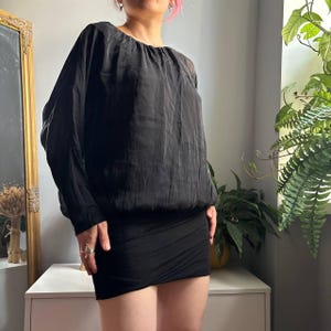 May include: A black long-sleeved blouse with a gathered round neckline and a loose, draped bodice. The garment features a fitted, solid black skirt section at the bottom, creating a two-tone appearance with a distinct contrast in fabric texture.
