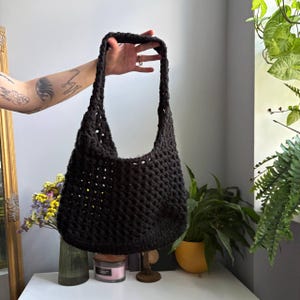 May include: A black crochet hobo bag featuring an open-work diamond pattern throughout the body and a wide, integrated shoulder strap. The bag is constructed from a thick, textured yarn and has a soft, slouchy silhouette with a rounded base.