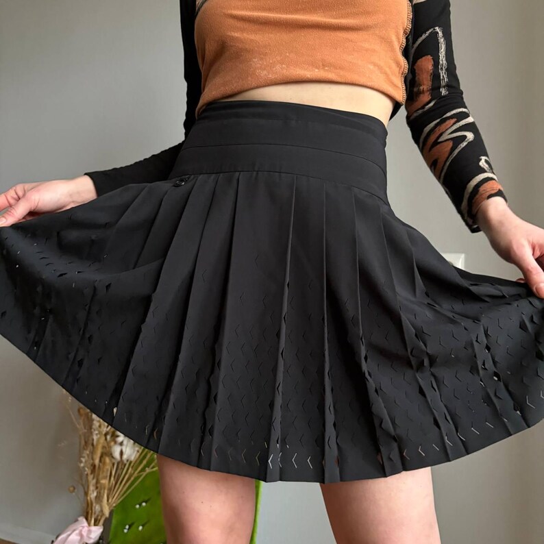 K&ouml;nnte beinhalten: Schwarzer Faltenrock mit dekorativen Ausschnittdetails. Der Rock hat eine hohe Taille und einen klassischen Tennisrock-Stil. Der Stoff scheint ein leichtes Material zu sein. Der Rock wird hochgehalten.