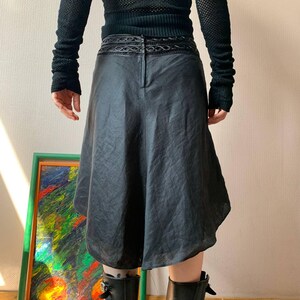 K&ouml;nnte beinhalten: Schwarzer, knielanger Rock mit einem einzigartigen, asymmetrischen Saum. Der Rock hat einen dekorativen Bund und einen dezenten Glanz. Er wird mit schwarzen Stiefeln und einem schwarzen Oberteil kombiniert, was einen monochromen Look ergibt.