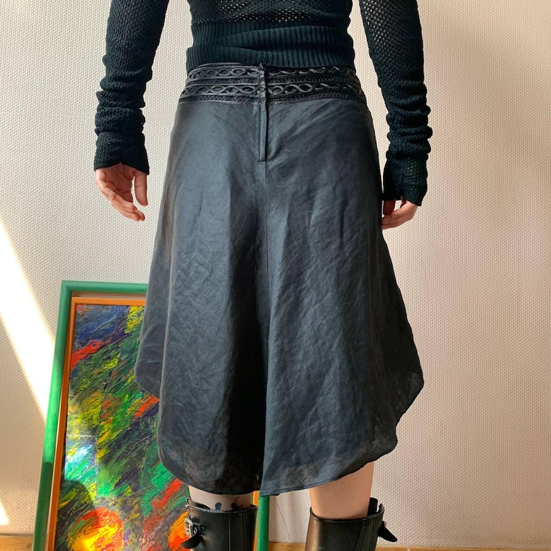 K&ouml;nnte beinhalten: Schwarzer, knielanger Rock mit einem einzigartigen, asymmetrischen Saum. Der Rock hat einen dekorativen Bund und einen dezenten Glanz. Er wird mit schwarzen Stiefeln und einem schwarzen Oberteil kombiniert, was einen monochromen Look ergibt.