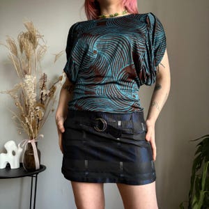 May include: A teal and brown patterned top with short sleeves and a black mini skirt with a belt. The top has a wavy design. The skirt has a grid-like pattern and a silver buckle.