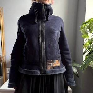 May include: A dark navy blue jacket with a high collar, a full-length front zipper, and adjustable buckle straps at the neck. The jacket features a subtle tonal floral pattern, dark trim along the zipper, and a rectangular patch of metallic gold and silver sequins on the lower front hem.