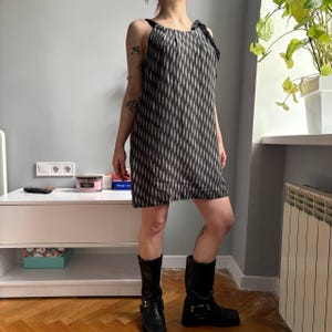 May include: A sleeveless, knee-length dress with a black and cream diamond pattern. The dress has black straps and a bow detail on one shoulder. The person is wearing black boots.