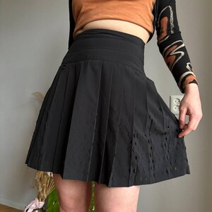 K&ouml;nnte beinhalten: Schwarzer Faltenrock mit hoher Taille und rautenf&ouml;rmigen Aussparungen am Saum. Der Rock ist aus einem glatten, matten Stoff gefertigt und hat eine klassische A-Linien-Silhouette. Der Rock ist ein vielseitiges Kleidungsst&uuml;ck f&uuml;r jede Garderobe.