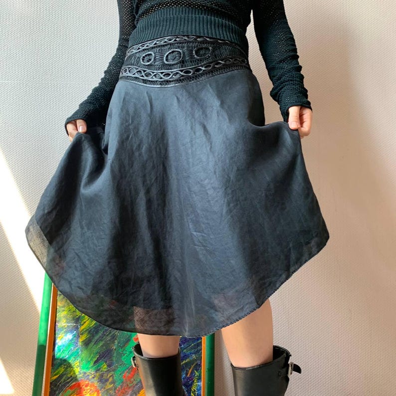 K&ouml;nnte beinhalten: Ein dunkelgrauer Rock mit dekorativem Bund. Der Rock hat eine ausgestellte Silhouette und einen transparenten Saum. Der Bund weist ein kompliziertes Design mit kreisf&ouml;rmigen und geometrischen Mustern auf. Die Person im Bild tr&auml;gt ein schwarzes Langarm-Oberteil und schwarze Stiefel.