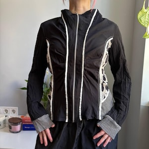 May include: Black jacket with white trim and a full-length zipper. The jacket features a high collar and long sleeves with striped cuffs. The back of the jacket has a patterned design in shades of white and beige.