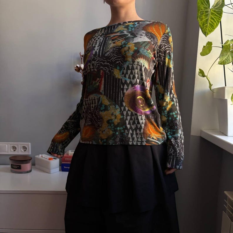 Puede incluir: Top de manga larga con un estampado abstracto colorido en tonos naranja, turquesa, morado y negro. Se combina con una falda negra. El dise&ntilde;o presenta formas geom&eacute;tricas y motivos florales.