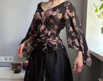 May include: A black sheer blouse featuring a vibrant floral and paisley print in shades of pink, yellow, and green. The top has a V-neckline, long flared sleeves, and a gathered front panel with adjustable drawstring ties at the waist.