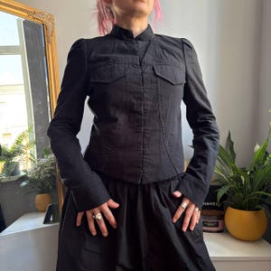 May include: A black long-sleeved jacket featuring a mandarin collar, a front hook-and-eye closure, and two chest pockets with flaps. The jacket has structured shoulders and a textured fabric finish, paired with a matching black skirt.
