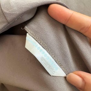 May include: A taupe-colored garment featuring a white fabric tag sewn into the interior seam. The tag displays light blue decorative stitching along the top edge and includes the text "amerie" printed in a clear, dark font on the white surface.
