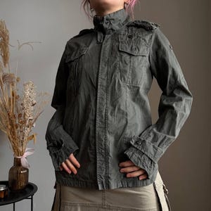 May include: Olive green military-style jacket with multiple pockets and button closures. The jacket features a high collar and epaulettes on the shoulders. The sleeves have buttoned cuffs. The jacket is made of a durable fabric.
